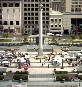 The Dewey Monument rises in the center of a busy urban plaza with white tents and people below.
