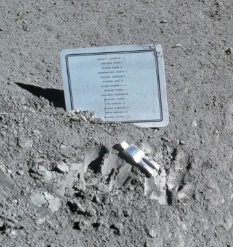 A small fallen astronaut figure lies on the lunar surface beside a memorial plaque.
