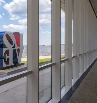 The Love sculpture is seen outdoors through large museum windows beside the water under a cloudy sky.