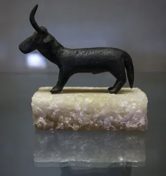 A dark Nuragic bronze statuette of a horned animal on a light rectangular base, shown with a reflected image on a glossy surface.