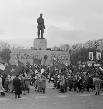 Crowd parades past the Stalin Monument, with flags, balloons, and portrait banners around the towering statue.