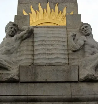 Relief figures on the Titanic Engine Room Heroes Memorial, showing two shirtless male figures beside a central panel with wavy lines and a sunburst above.