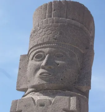 Close-up of an Atlantean figure at Tula with a carved face and patterned headdress against a blue sky.