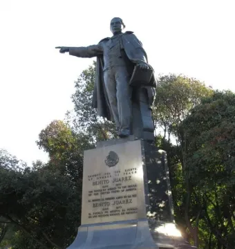Bronze statue of Benito Juarez standing on a multi-tiered stone pedestal, holding papers and pointing forward.