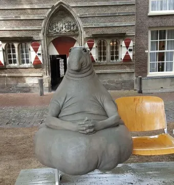 Homunculus Loxodontus sculpture seated on a metal bench outside, with a brick building behind it.