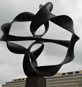 Continuum sculpture with twisting black metal ribbons on a tall pedestal against an overcast sky.