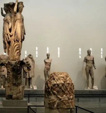 Dancers of Delphi sculpture with three draped female figures on a carved column base in a museum hall.