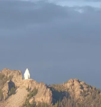 Our Lady of the Rockies statue on a rocky mountain peak beneath dark clouds, with a communications tower on a nearby ridge.