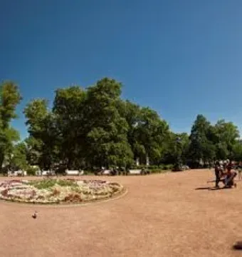 Silhouetted Catherine II Monument in a sunlit square with trees, historic buildings, and people walking nearby.