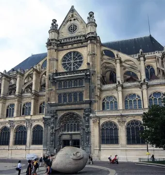 Écoute, a pale stone head sculpture with a hand cupped to its ear, stands in a plaza with a church behind it.
