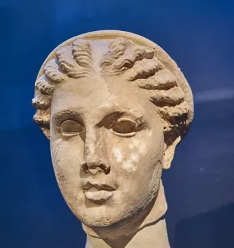 Weathered stone head of Lycosoura Artemis with braided hair on a small pedestal.