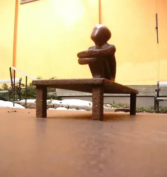 Järnpojke, a small dark seated sculpture on a low bench beside a building wall with patches of snow and greenery.