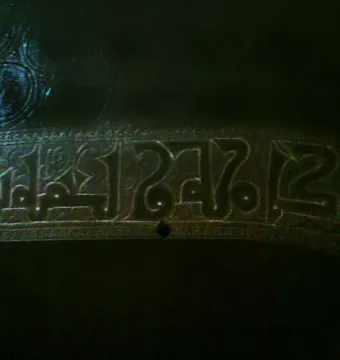 Close-up of the Pisa Griffin showing a band of raised Kufic calligraphy and circular motifs on the bronze surface.