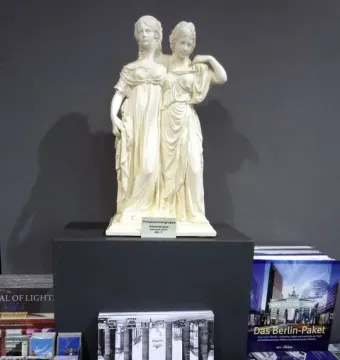 Small white Princesses Monument statuette of two women in classical dresses, displayed on a pedestal among books and souvenirs.