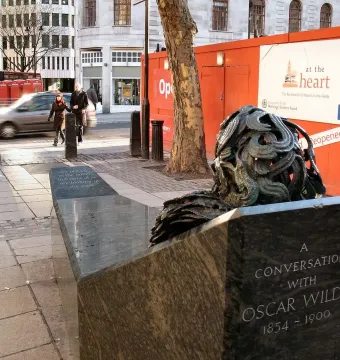 A Conversation with Oscar Wilde bench sculpture with an abstract bronze-toned bust and swirling hair on a city sidewalk.