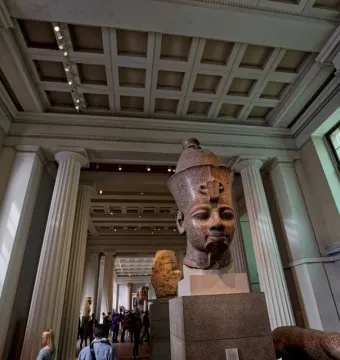 Colossal head of Amenhotep III displayed on a pedestal in a museum gallery, with a large stone fragment beside it.