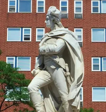 Marble statue of George Washington in period attire and a cape, shown beside a brick building.