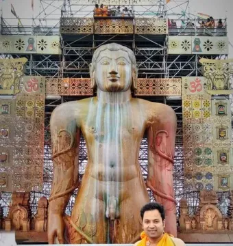 Gommateshwara Bahubali statue with ritual streaks, framed by a decorated scaffolding structure and a smiling man in orange robes in the foreground.