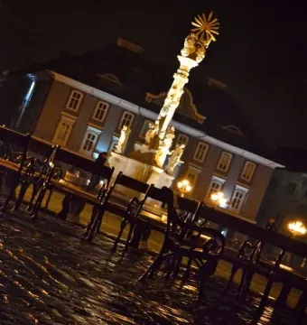 Holy Trinity Monument illuminated at night above wet cobblestones and park benches