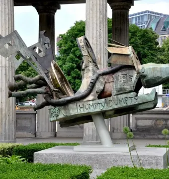 Humpty Dumpty Machine bronze sculpture with a green and brown patina, shown outdoors on a plinth beside classical columns.