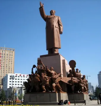 Colossal bronze statue of Mao Zedong with his right arm raised, standing above a multi-tiered base with bronze figures below it.