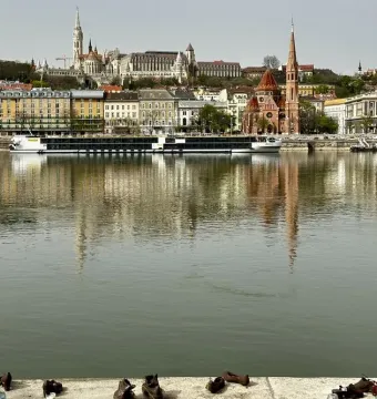 Shoes on the Danube Bank memorial along a riverside embankment, with a city skyline and riverboat in the background.