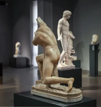 The Subiaco Ephebe sculpture on a dark pedestal in a museum gallery, with other classical statues in the background.