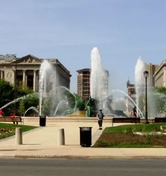Swann Memorial Fountain in a city plaza, surrounded by paved walkways, trees, and classical stone buildings.