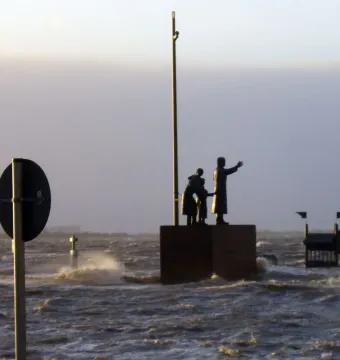 Auswandererdenkmal standing in choppy floodwater under an overcast sky, with a dark circular sign in the foreground.