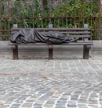 Homeless Jesus sculpture showing a sleeping figure covered by a blanket on a park bench, with wounded feet visible.