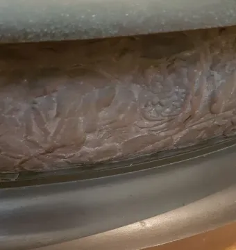 Close-up of Le Miroir showing a dark sculpted base with intricate relief patterns and a rounded edge resting on a wooden surface.