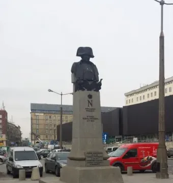 The Emperor Napoleon I Memorial, a dark bust of Napoleon in a bicorne hat on a tall stone plinth in a city square.