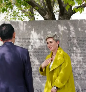 Three people stand in front of the Japanese American Memorial to Patriotism During World War II, beside a stone wall engraved with names under leafy tree branches.