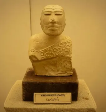 Bust of the Priest King sculpture with a decorative headband and patterned cloak on a dark base