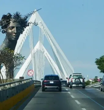 Reminiscencia, a dark bust of a woman crowned with flowers and spiky elements, set before a white cable-stayed bridge.