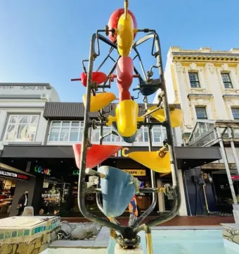 The Bucket Fountain sculpture with colorful buckets in a light blue basin on Cuba Mall, with buildings and shop fronts behind it.