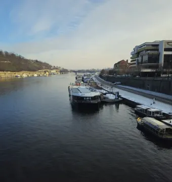 Winter view of the Vltava River with moored boats, a snowy embankment, and a wooded hill across the water; the Prague Metronome is not clearly visible.