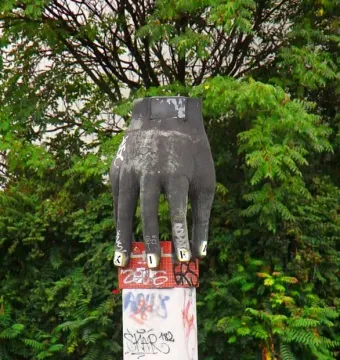 Weathered Hand with Clock sculpture standing on a tiled pedestal with graffiti, framed by trees and green foliage.