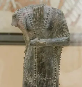 Iddi-Ilum statuette of a headless standing figure with folded arms and a decorated robe on a square base.