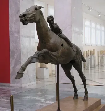 The Jockey of Artemision bronze statue displayed on a pedestal in a museum hall, with the horse rearing and a small rider on its back.