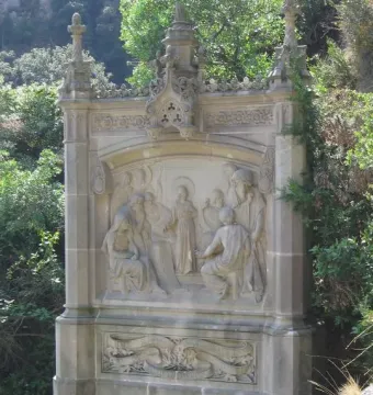 Ornate stone relief on the Monumental Rosary of Montserrat showing several figures gathered around a young person, with decorative architectural details and carved banners.