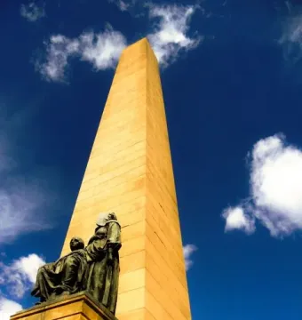 National Women\'s Monument with a tall obelisk and bronze figures of two women on the pedestal against a blue sky.