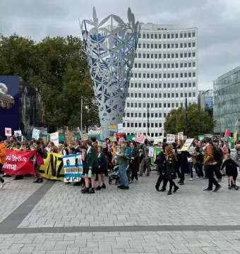 The Chalice sculpture in a public square with a crowd gathered around its base, carrying colorful banners and signs.