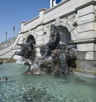 Court of Neptune Fountain with dark metal sea deities and horses rising from a water pool beside an ornate stone building.