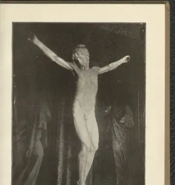 Black and white photograph of a crucifix sculpture showing a nude bearded figure with outstretched arms against a dark background.