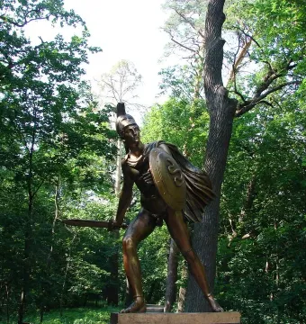 Bronze Greek Warrior Monument of an armored fighter with a shield and raised sword in a forest setting.