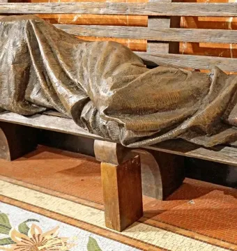Bronze Homeless Jesus sculpture of a covered figure lying on a slatted bench, with bare feet visible at one end.