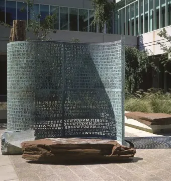 Kryptos bronze sculpture covered with carved capital letters, standing outdoors near stone benches and trees.