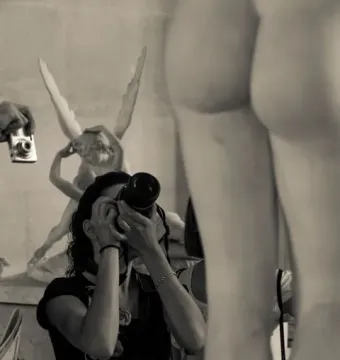 A museum visitor photographs a marble winged sculpture, with another large sculpture blurred in the foreground.