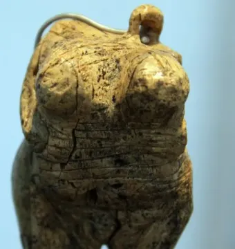 Venus of Hohle Fels figurine with cracked ivory torso, prominent breasts, and a loop at the top on a dark stand.